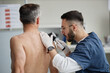 © Seventyfour - Middle aged Caucasian man sitting with back exposed while young adult male dermatologist examining skin on shoulder using dermatoscope during medical consultation