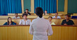 © Raushan_films - Indian woman teacher teach young adult male female sitting desk table do study learn new course exam indoor class room. Female tutor talk speak share idea plan teen group attend college day