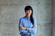 © bnenin - Asian Woman in Blue Shirt Stands Confidently With Crossed Arms in Modern Workspace