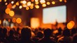 © Iryna - Rear view of crowd sitting in dark auditorium listening to speaker with blurred warm orange bokeh lights and large projection screen conference seminar or live event atmosphere