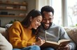 © Vadym - Happy multiracial couple shares book and laughs together on couch. Woman writes in notebook while man reads, enjoying cozy home time during cold season.