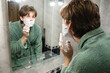 © svetlana_cherruty - Young man standing at the bathroom mirror and spreading thick shaving foam on his face before shaving as part of daily grooming routine. Morning Routine. Side view.