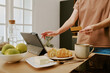 © AnnaStills - Caucasian young adult woman holding credit card and using tablet for online shopping in kitchen, left hand touching screen while standing near plate with croissants and cup of coffee