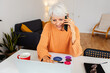 © Xavier Lorenzo - Smiling senior business woman working on laptop from home, talking on the phone and taking notes on a business plan. Entrepreneur people and business concept