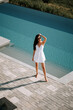 © Darius - Woman in white dress enjoying sunny day by clear swimming pool in luxurious setting
