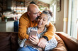 © Geber86 - Mature husband and wife smiling at smartphone on sofa at home