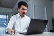 © fotolite - Young man focuses on his laptop while working from home. Clean indoor setup showing concentration and modern remote work.