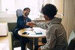 © Maskot - Curly haired teenage boy shaking hands with male counselor while sitting in high school office