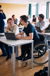 © Maskot - Young man sitting on wheelchair talking with male classmate while studying on laptop in high school classroom
