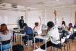 © Maskot - Rear view of male students raising hand for asking questions to teacher in classroom at high school