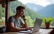 © Gabriel - Young man work on laptop and talk on phone while sitting in outdoors cafe in mountain resort during summer vacation. alternative office, nomad remote work concept, business online using Internet