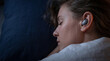 © Marif - Serene young woman peacefully sleeping in a dark bedroom, wearing wireless earbuds to listen to calming music or a podcast for deep relaxation and rest