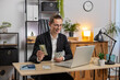 © Andrii Iemelianenko - Happy young businessman counting dollar money cash and using laptop computer in home office. Successful freelancer guy getting money in office, calculates dividends cash profits and finances at table.