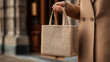 © Maryna - A close-up of a person's hand holding a modest burlap bag while dressed in a stylish beige coat, set against an urban backdrop.