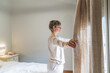 © Koldo_Studio - Woman standing in pajamas, opening bedroom curtain, inviting morning light and new beginnings into her home space