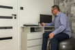 © dominikspalek.pl - Male office worker in blue shirt sitting on orthopedic ball in front of computer, office workers back problems
