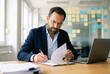 © Facundo - Concentrated businessman with glasses signing a contract paper at a desk with a laptop in a modern office