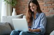 © miss irine - Young woman smiles using laptop on couch. She browses internet in living room, works remotely. Female freelancer happy with wireless device at home.
