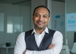 © Yannick - Smiling Indian businessman with arms crossed, sharp corporate vest, glass office