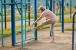 © Татьяна Яблокова - Full body side view of an 87-year-old woman performing a hamstring stretch, touching her foot to a bar on a blue climbing ladder at an outdoor fitness area. Flexibility for seniors.