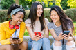 © Xavier Lorenzo - Three cheerful young women are enjoying a summer day outdoors, sitting together and sharing a laugh while using their smartphones