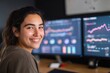 © sogap - A smiling Latina woman confidently working with financial data on dual computer screens at her desk