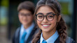 © sommersby - Young, happy female student with glasses and braided hair smiles brightly, with another blurred student in uniform visible behind her, embodying joy of school life