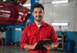 © Vasiliy - Confident auto mechanic in a red uniform smiling at the camera in a repair shop. Professional technician holding a clipboard with a car on a lift in the background