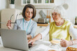 © shurkin_son - Social support for seniors easing tech access, female social worker teaching retired lady how to use laptop and online applications, both sitting in cozy kitchen at table and having tea