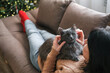 © Elen Nika - Woman in cozy sweater is relaxing on sofa petting fluffy gray cat on her chest with decorated Christmas tree in blurred background. Christmas comfort, pet love, festive season, relaxing winter weekend
