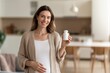 © Saniya - Smiling pregnant woman holding a white pill bottle, symbolizing prenatal care, health, and the joy of expectant motherhood in a bright home.