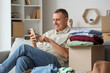 © Pixel-Shot - Mature man using mobile phone while unpacking box with clothes at home