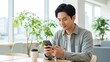 © Watu ireng - Young Asian man smiling while using smartphone in a bright modern office with plants.