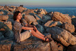 © soleg - A girl poses and relaxes by the sea, sitting on rocks near the slope of a high cliff, panoramic view of the seashore and mountains at sunset