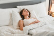 © artitwpd - A young woman rests peacefully on a bed with white sheets in a bright, cozy bedroom, capturing a serene moment of sleep, relaxation, and wellness in a calm, supportive home environment.