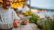 © Pinklife - A mature man with a gray beard sits at a rustic wooden table, intently reading on his tablet surrounded by flowers, conveying a feeling of tranquility and contemplation in a café setting.