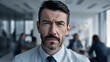 © Yuliia - Serious businessman with salt and pepper beard in white shirt looking intensely at camera, office environment with blurred colleagues in background.