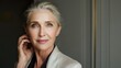 © Yuliia - Elegant mature woman with silver hair in light beige outfit against neutral background, touching face gently with thoughtful expression.