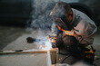 © qunica.com - A skilled welder crouches beside a metal frame, sparks and smoke rising as they fuse joints. Wearing a protective helmet and gloves, the focused worker performs a precise weld in a workshop setting.