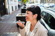 © Marko Geber - Adult woman smiling using voice assistant on city street