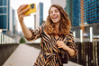 © maxbelchenko - Selfie time! Young woman is enjoying herself with phone in bustling cityscape. Beautiful tourist enjoys stroll, blogs on sunny day against backdrop of metropolis. Blogging, urban, tourism concepts.