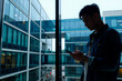© pressmaster - Young adult Asian man standing by large window using smartphone, modern office building exterior visible through glass, subject focused on device, casual posture, urban setting