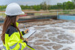 © reewungjunerr - Professional Asian female environmental engineer conducting an inspection and recording operational data on a clipboard at a large industrial water treatment plant sewage facility outdoors.