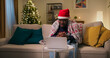 © ABCreative - A cheerful young man of multi-ethnic background sits on a sofa in the living room, having a video chat on his laptop. Covered with a blanket and wearing a Santa hat, he communicates and smiles.