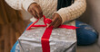 © ABCreative - Preparing gifts for friends, a young woman of multi-racial background holds a gift in a close-up. Dressed in jeans and a warm white sweater, she carefully ties a red ribbon on the bow.