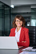 © Stock 4 You - Vertical portrait middle-aged specialist latin hispanic business lady working on laptop pc sitting at desk in office smiling at camera. Mature CEO woman in red suit using computer technology for work