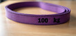 © Pete - Purple lifting sling marked 100 kg rests on wood surface. Heavy duty industrial strap for cargo, safe load lifting. Rigging equipment, construction safety tool.