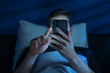 © Arsenii - Close up of young man, alone in the dark bedroom, holds his smartphone close to face while scrolling social media or texting late at night. Screen dependence, excessive consumption of content