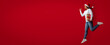 © Prostock-studio - A person wearing a Santa hat jumps happily while holding a gift on a vibrant red background. The scene captures the joy of the holiday season and festive spirit.