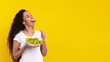 © Prostock-studio - A young woman with long curly hair is holding a bowl of fresh salad and smiling as she takes a bite of lettuce. The bright yellow background enhances her joyful expression and healthy meal choice.